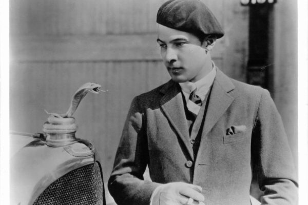 Rudolph Valentino is looking at a snake radiator mascot on hood of car, 1925.